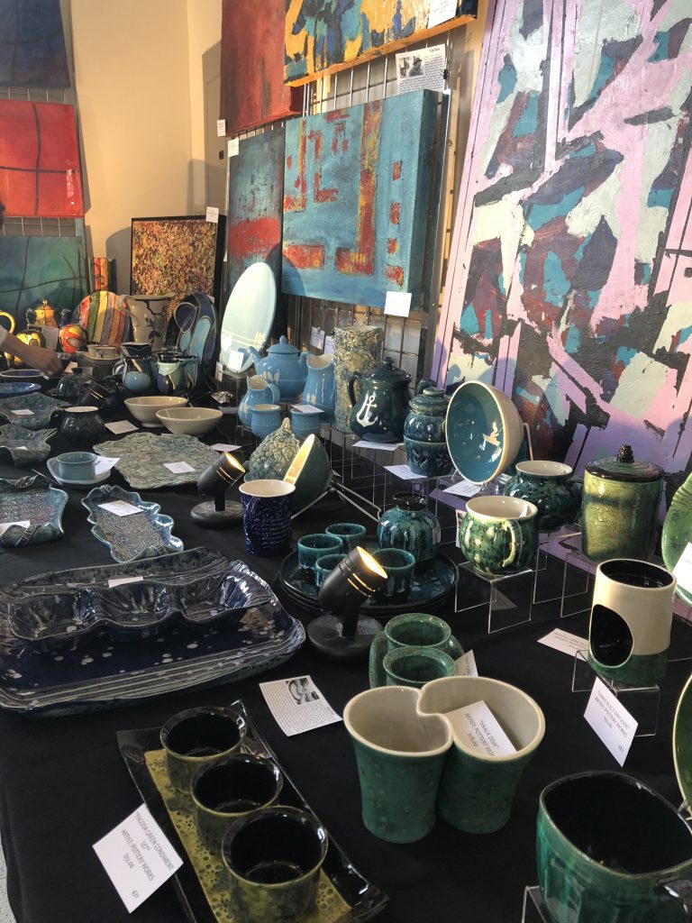 A table of blue and green ceramic artwork including bowls and mugs with a rack of abstract paintings hung up behind it.
