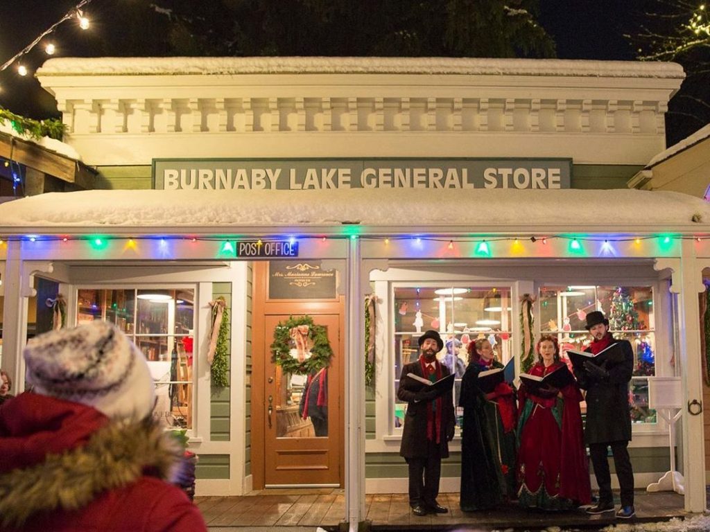A group of four carollers in early 20th century festive clothing sing for a crowd in front of the Burnaby Lake General Store in the Heritage Village.