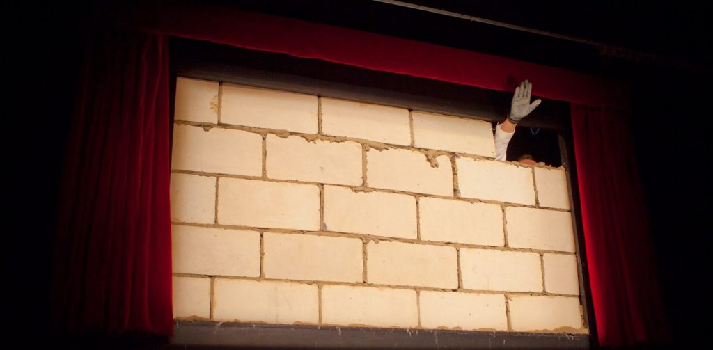 A hand salutes through a hole in a brick wall during Free Admission performance