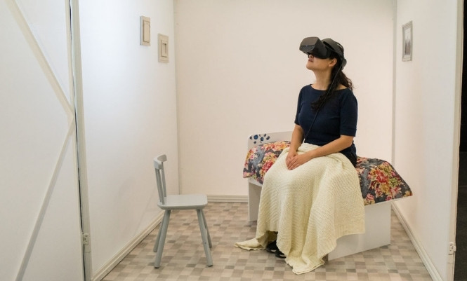 W woman wearing a VR headset sits on a bed in a small, sparse, white room.